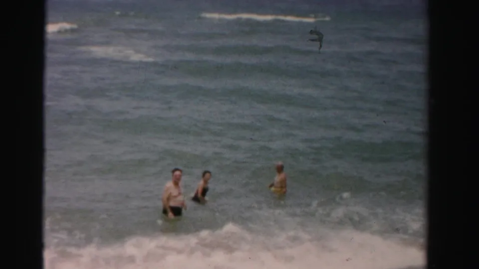 Adults in swimwear wade separately in shallow ocean water in 1955 Dade County footage, with a broad view of waves and film-frame edges.