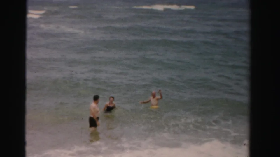 Three adults wade in the Dade County ocean surf, with one person gesturing and low waves rolling toward shore.