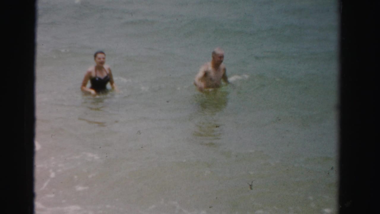 Two adults stand waist-deep in shallow ocean water near small waves.