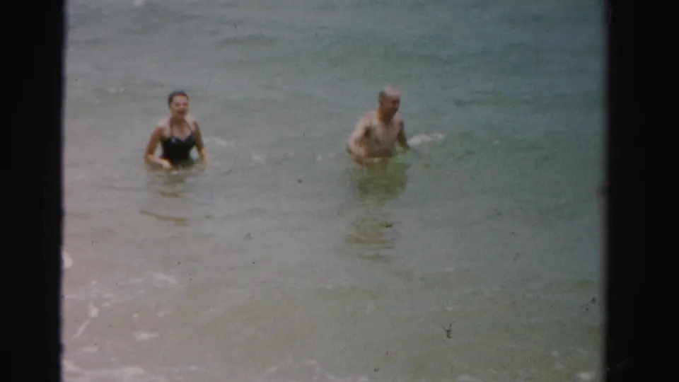 Two adults wade in shallow surf in 1955 Dade County, Florida, with the woman in a dark one-piece swimsuit and the man shirtless.