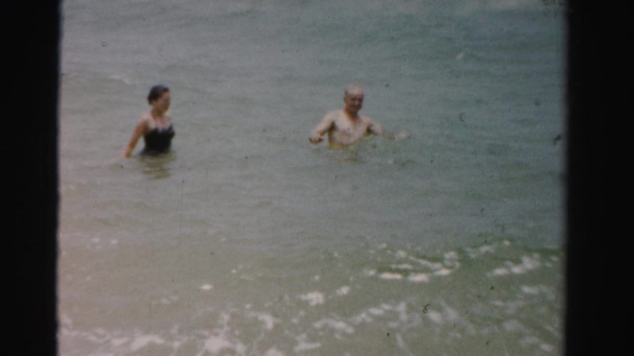 A woman and man stand apart in shallow surf with open water behind them.