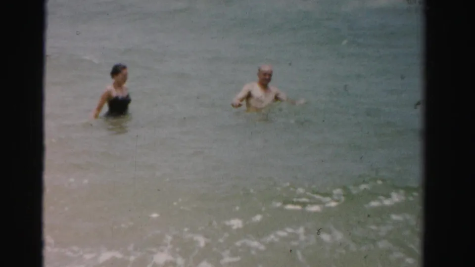 A woman in a dark swimsuit and a shirtless man stand in the ocean during a 1955 Florida home-movie beach scene.