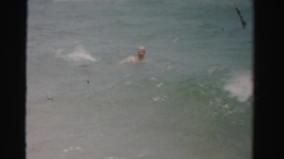 The man is partly submerged in the ocean while a small wave breaks across the 1955 Florida beach home-movie frame.
