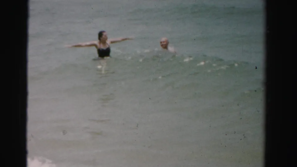 1955 Dade County home-movie frame with a woman in a dark swimsuit and a man in the surf.