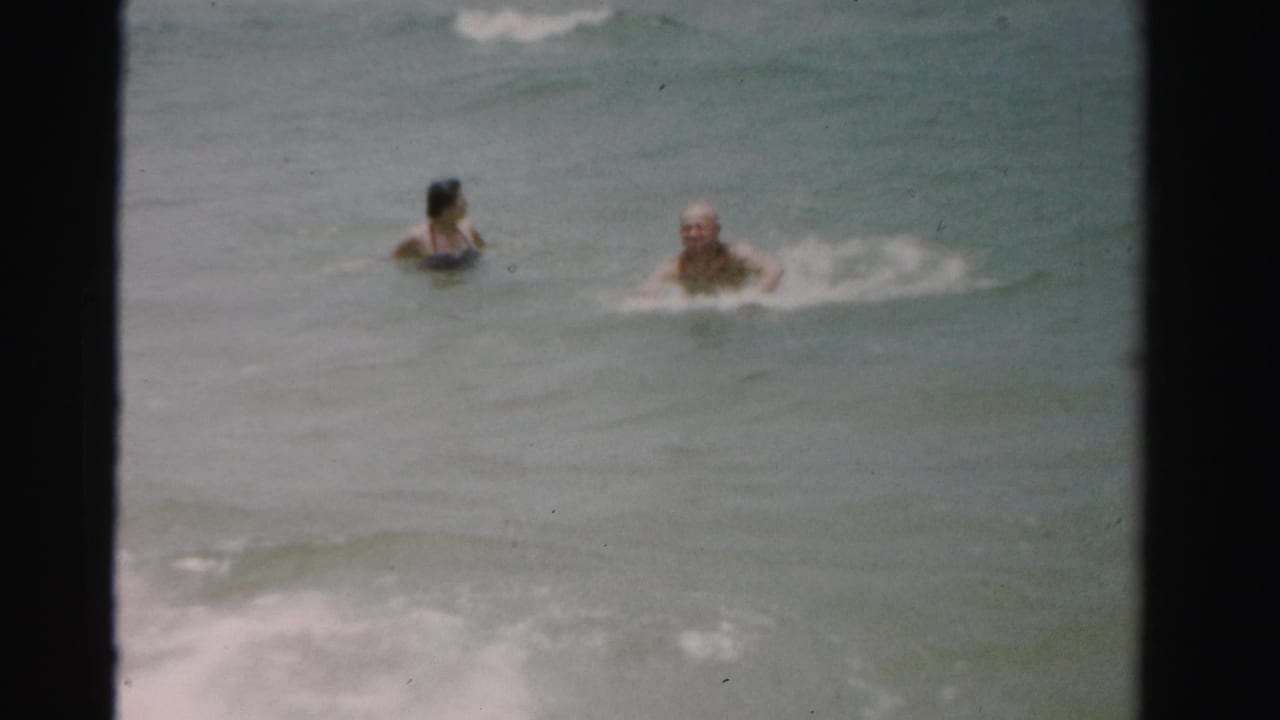 1955 Dade County 8mm frame showing two adults low in the water as a small wave breaks nearby.