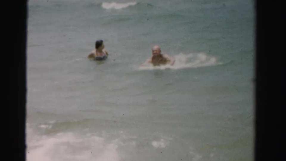 1955 Dade County 8mm frame showing two adults low in the water as a small wave breaks nearby.