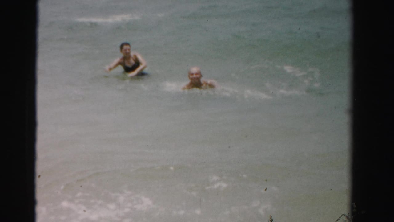1955 Dade County home-movie frame with two adults swimming toward the camera through gentle surf.