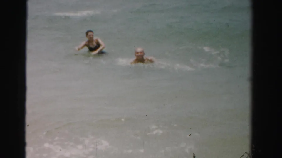 1955 Dade County home-movie frame with two adults swimming toward the camera through gentle surf.