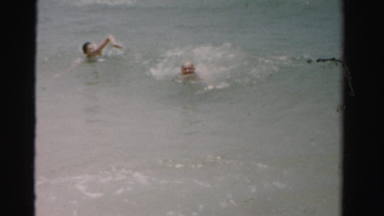 Two swimmers partly submerged in pale ocean water with surf around them.