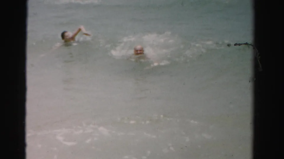 Two people swim in the ocean in 1955 Dade County, Florida, with soft 8mm grain and water splashing around them.