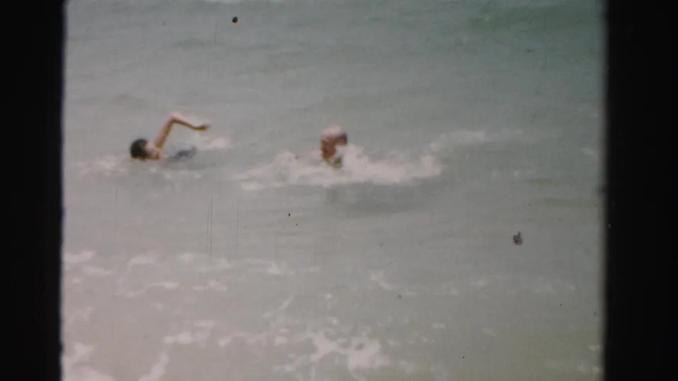 Two swimmers continue through the surf in 1955 Dade County, Florida, one raising an arm above the water.