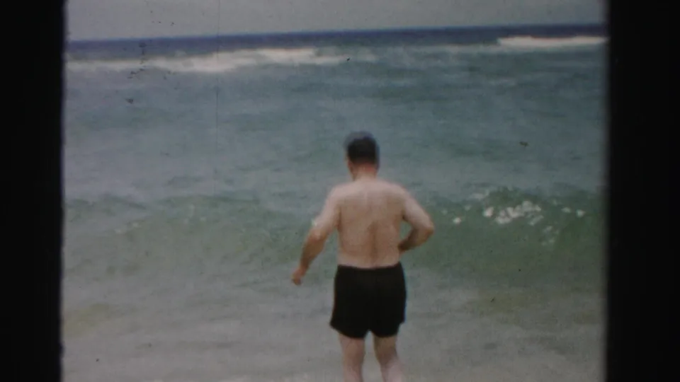 A man in dark swim trunks stands near the surf in 1955 Dade County, Florida, facing the ocean from the beach edge.