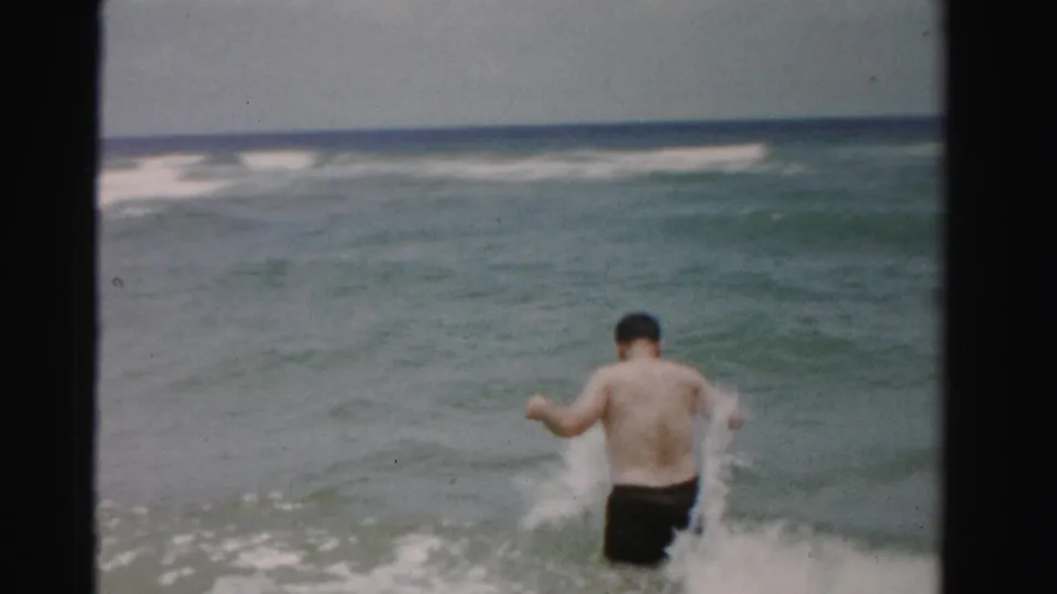 A man runs into the breaking surf in 1955 Dade County, Florida, seen from behind in grainy 8mm footage.
