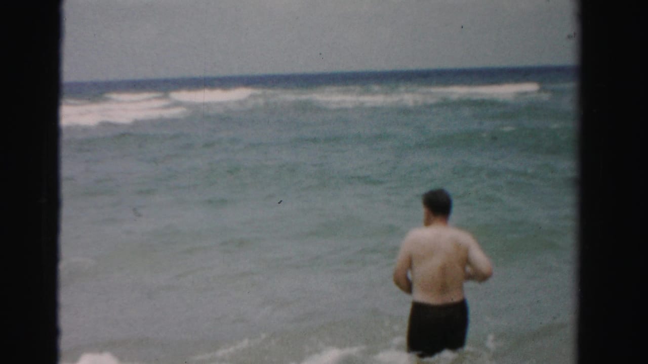 Adult man in dark swim trunks wading into ocean surf under a muted sky.