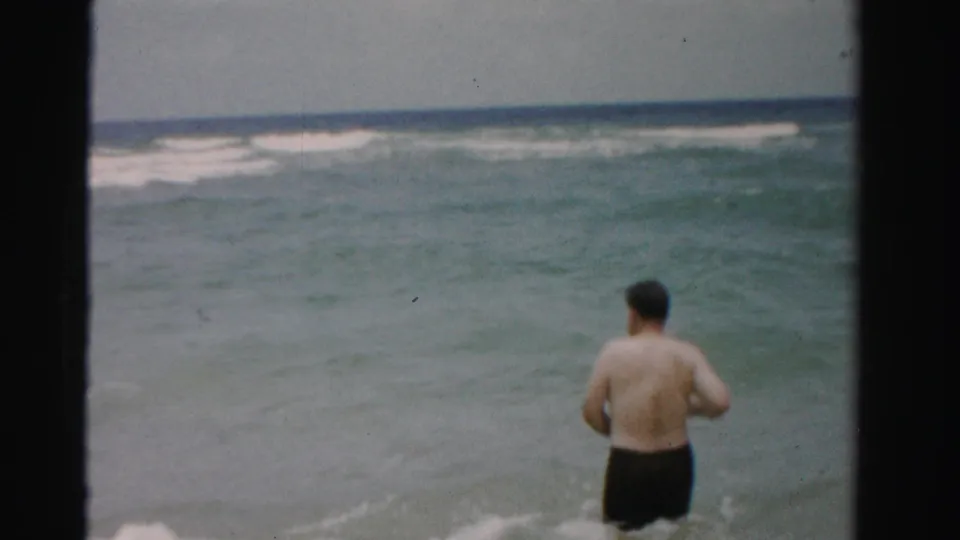 1955 Dade County home-movie frame showing an adult man from behind entering green-blue surf with whitecaps offshore.