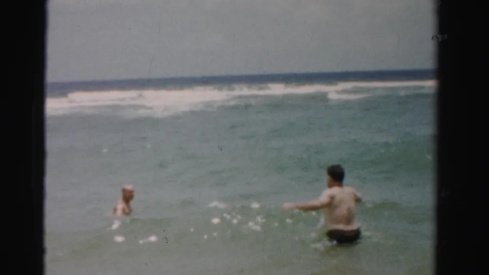 An 8mm home-movie frame from Dade County shows two adults in green-blue surf, one closer from behind and one farther left.