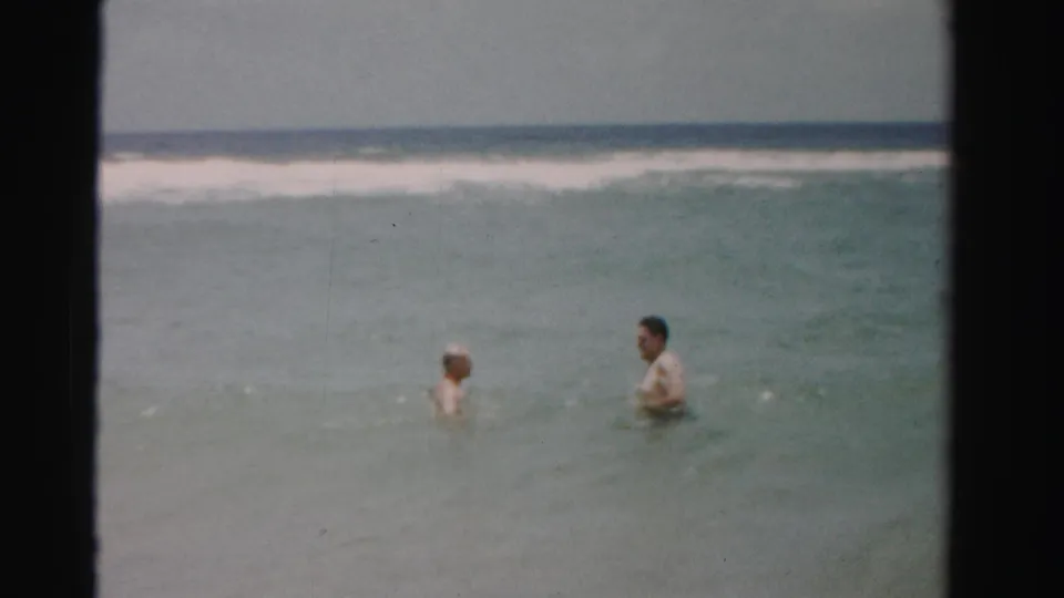 Two men remain in the shallow ocean, one seen in profile and one facing partly toward camera, in color 8mm footage.