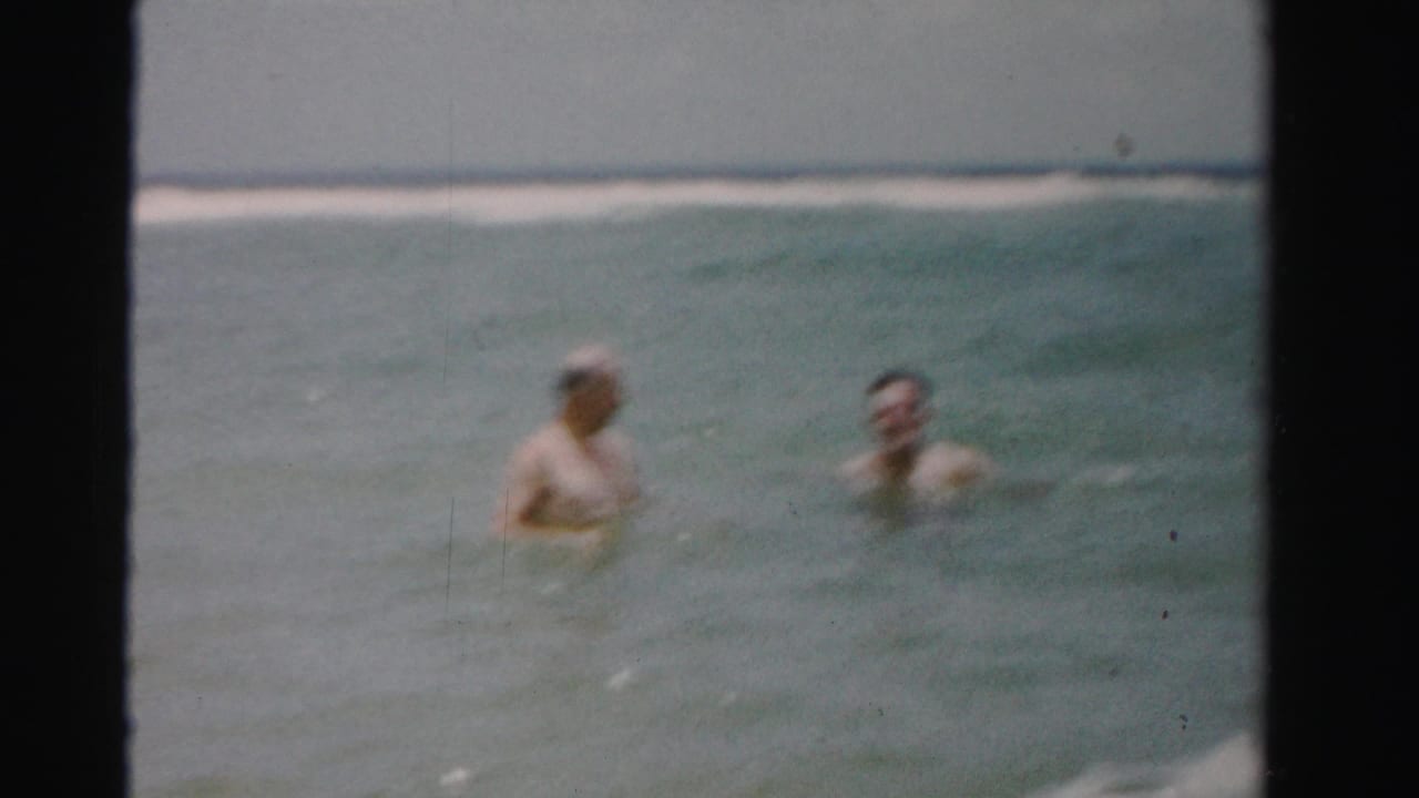 Two adult men wade in ocean water with a low surf line behind them.