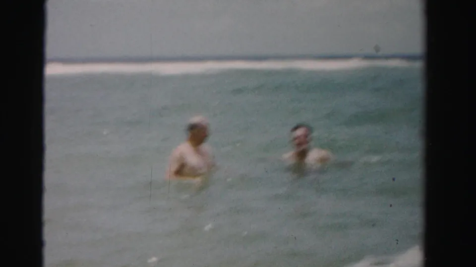 Two adult men wade chest-deep in the ocean in 1955 Dade County, Florida home-movie footage, with soft focus and dark film edges.