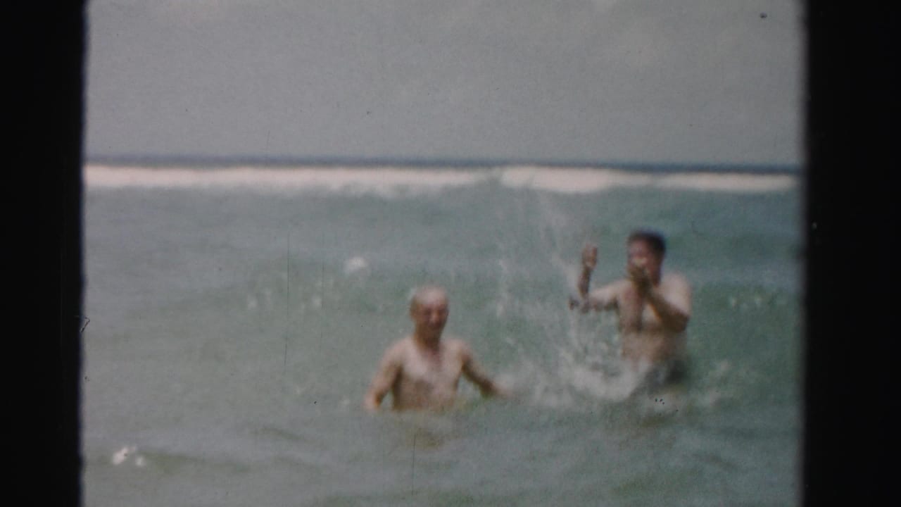 Two adult men stand in the surf as one splashes or waves toward the camera.