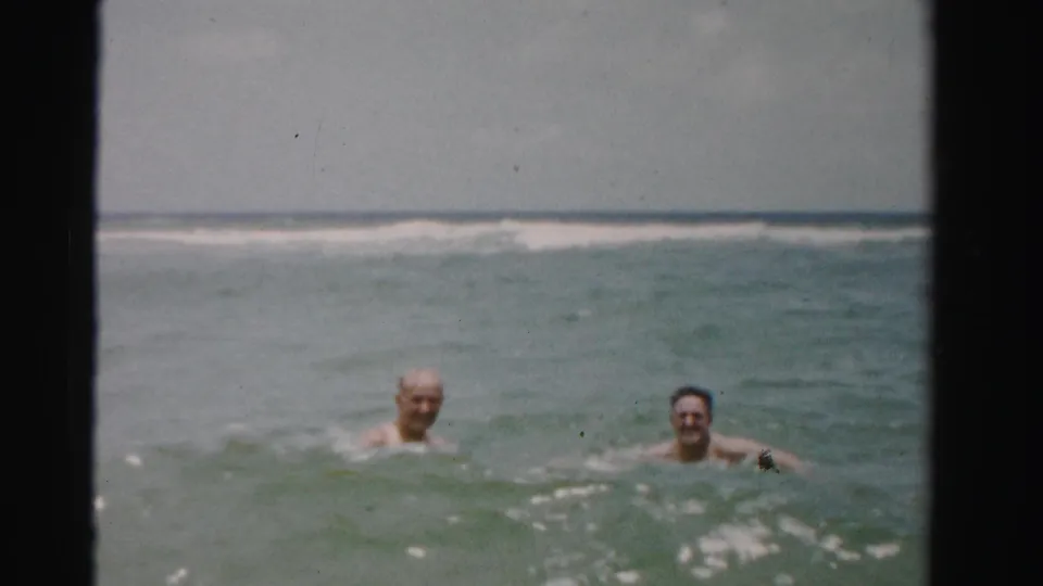 Two adult men appear to smile toward the camera while swimming in green-blue ocean water in 1955 Florida home-movie footage.