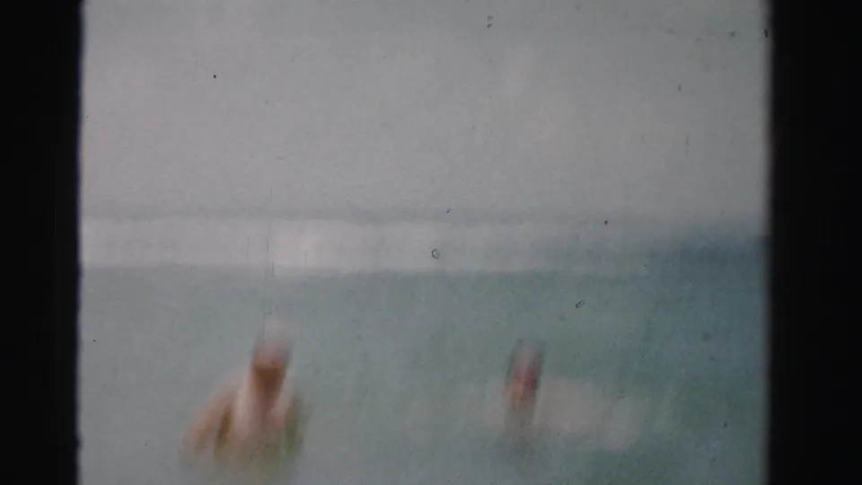 A soft, blurred 8mm frame shows two adult swimmers partly submerged in the ocean, with the horizon and surf faintly visible.