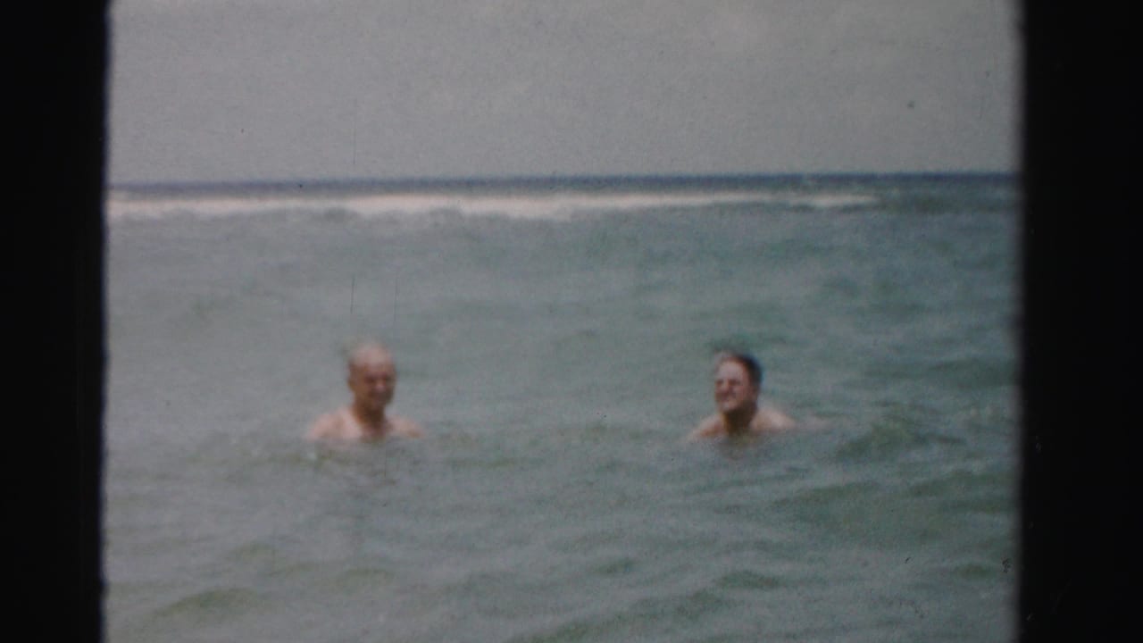 Two adult men swim neck-deep in ocean water with distant surf behind them.