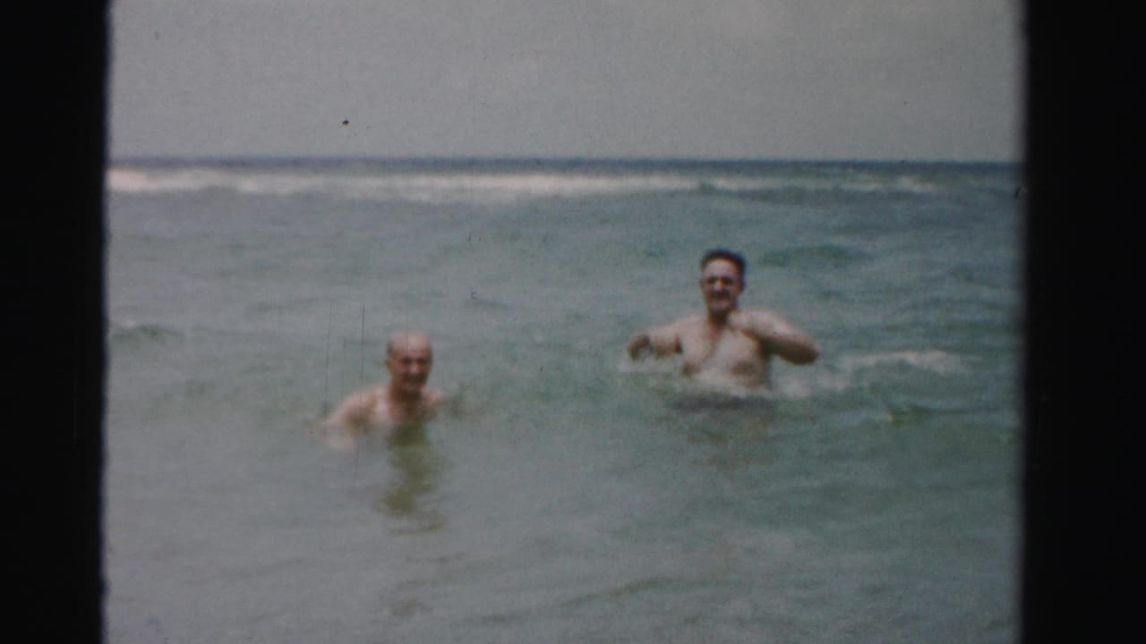 Two adult male swimmers face the camera from neck-deep water, with low surf and a hazy horizon in 1950s Dade County, Florida footage.