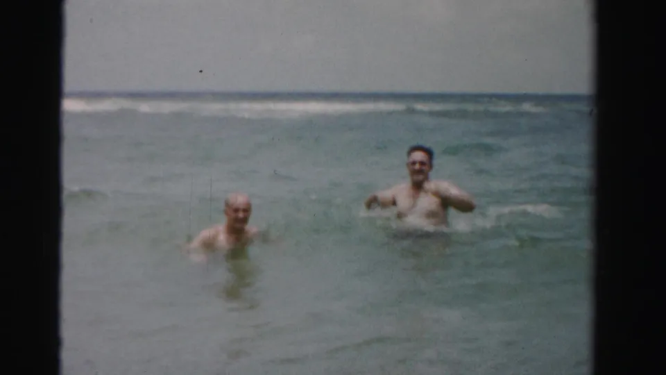 Two adult male swimmers face the camera from neck-deep water, with low surf and a hazy horizon in 1950s Dade County, Florida footage.