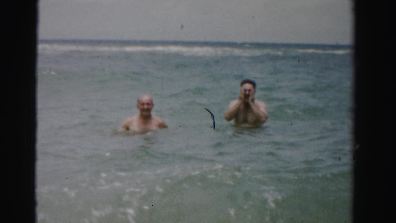 In the ocean off Dade County, Florida, two adult men remain chest- to neck-deep as the dark-haired man lifts his hands toward his face in a playful home-movie moment.