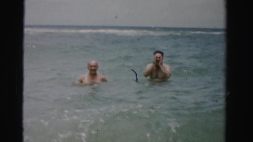 In the ocean off Dade County, Florida, two adult men remain chest- to neck-deep as the dark-haired man lifts his hands toward his face in a playful home-movie moment.