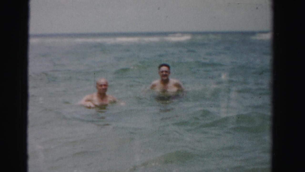 Two adult men smile while wading in gentle ocean surf, shown in faded 8mm home-movie footage identified as 1955 Dade County, Florida.
