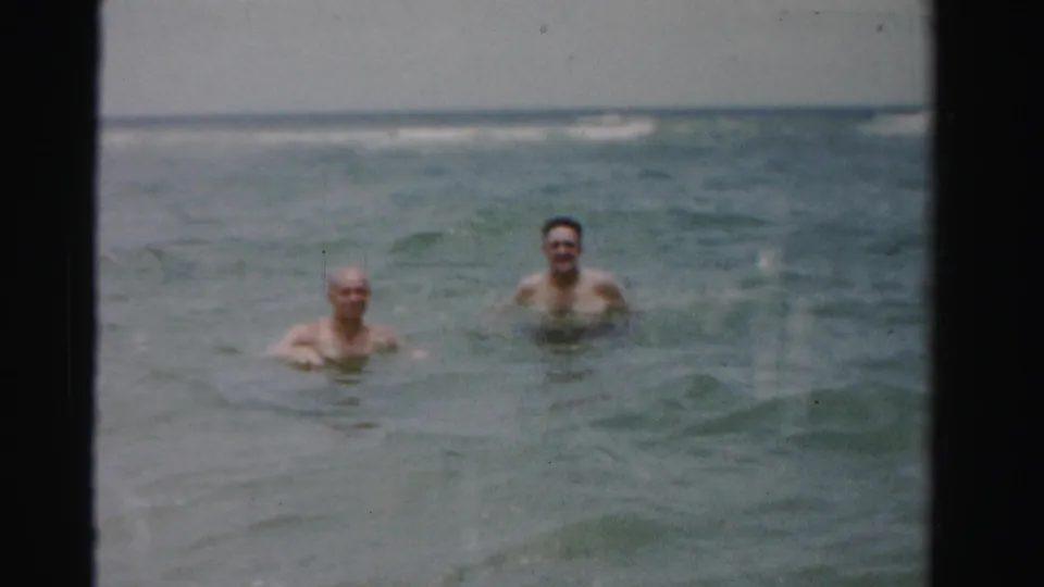 Two adult men smile while wading in gentle ocean surf, shown in faded 8mm home-movie footage identified as 1955 Dade County, Florida.