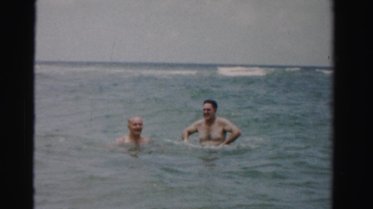 Two adult men stand chest-deep in ocean water with waves behind them.