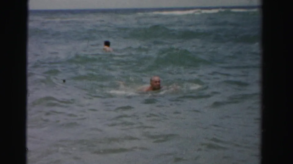 An older adult man moves through rough ocean water as another swimmer appears farther out in the surf.