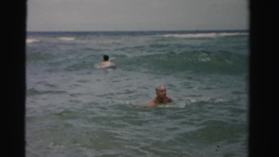 An older man remains near the camera as a second adult swimmer faces away in the Dade County surf.