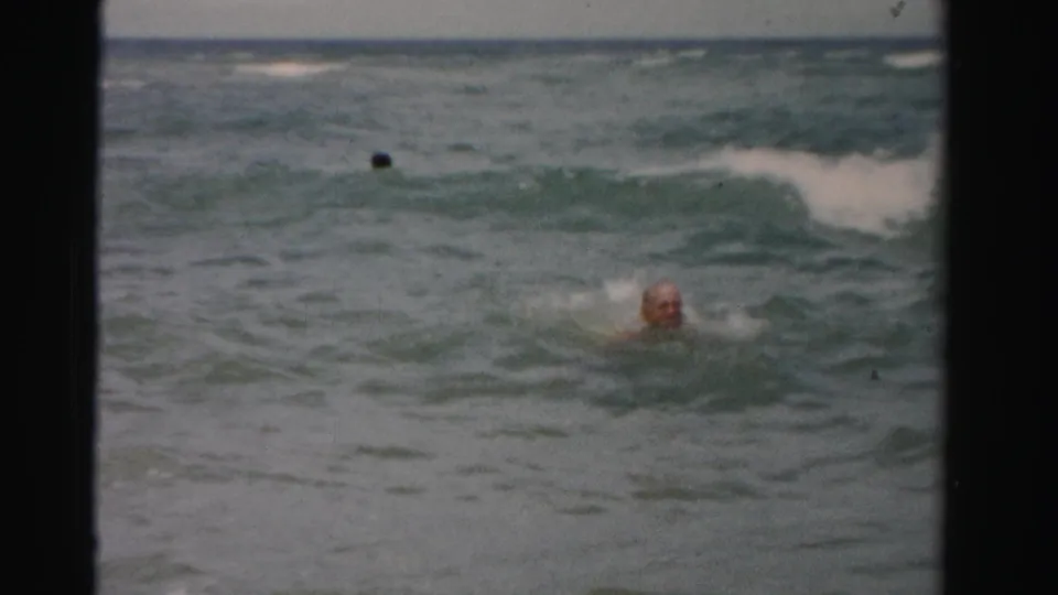 An older man swims through choppy ocean water as a second adult swimmer is partly visible beyond the wave line.