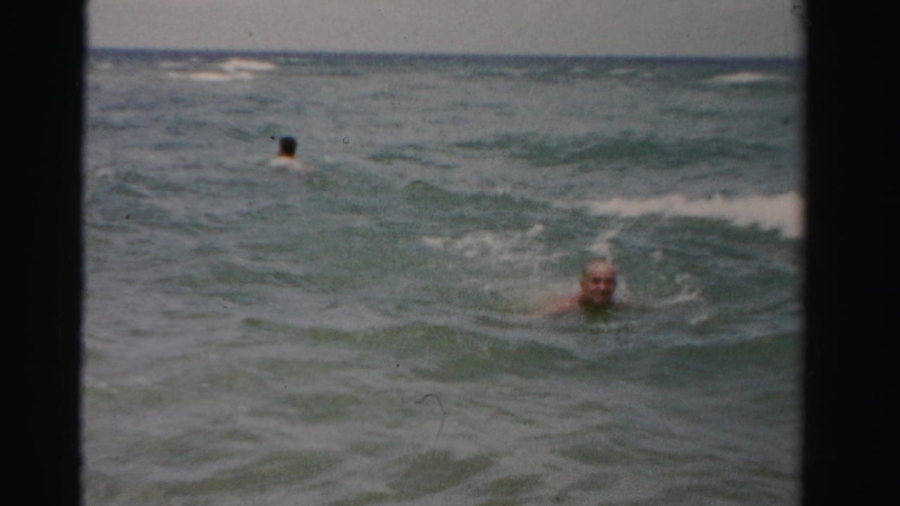 Older man swimming in ocean surf with another swimmer in the distance.