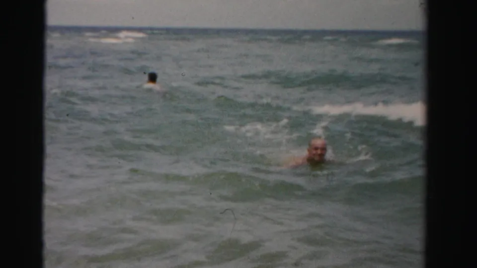 1955 Dade County home-movie frame showing an older man swimming through choppy ocean water with a distant swimmer behind him.