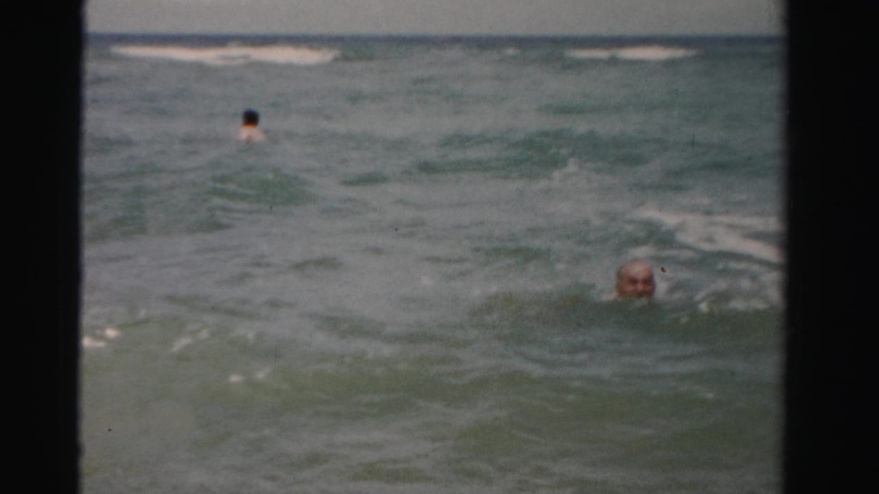 Older swimmer partly submerged in choppy ocean water near a distant swimmer.