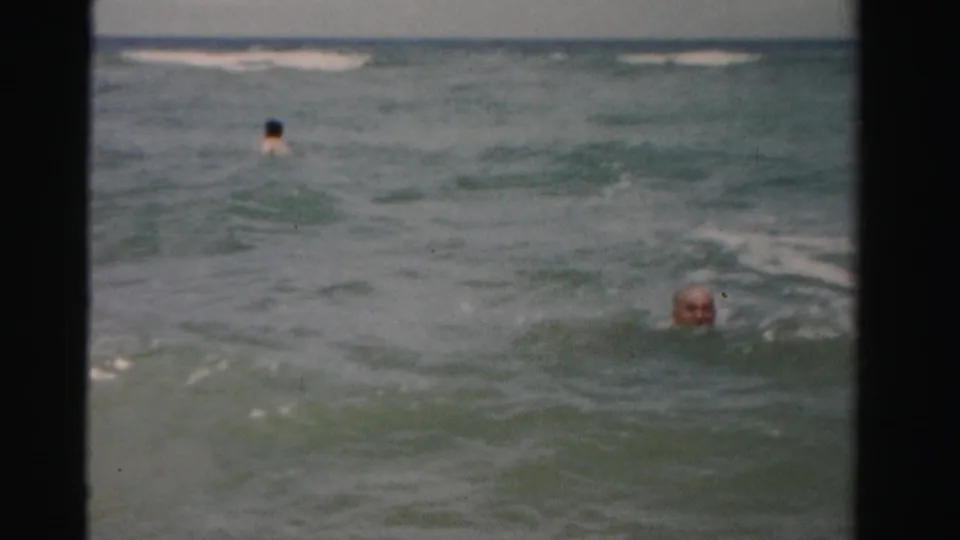 1955 Dade County frame with an older man partly visible in the surf and another swimmer farther out in the ocean.
