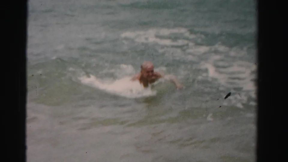 1955 Dade County home-movie still showing an older man closer to shore, pushing through foamy ocean surf.