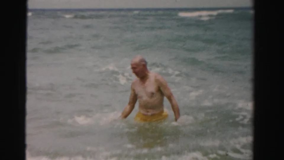 1955 Dade County frame showing an older man wearing yellow swim trunks as he wades through shallow surf toward shore.