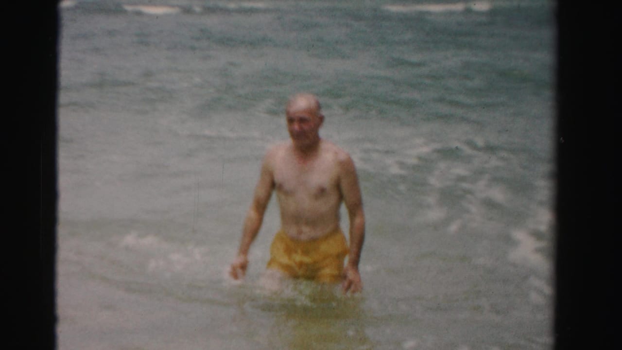 Older man in yellow swim trunks wading in shallow ocean surf.