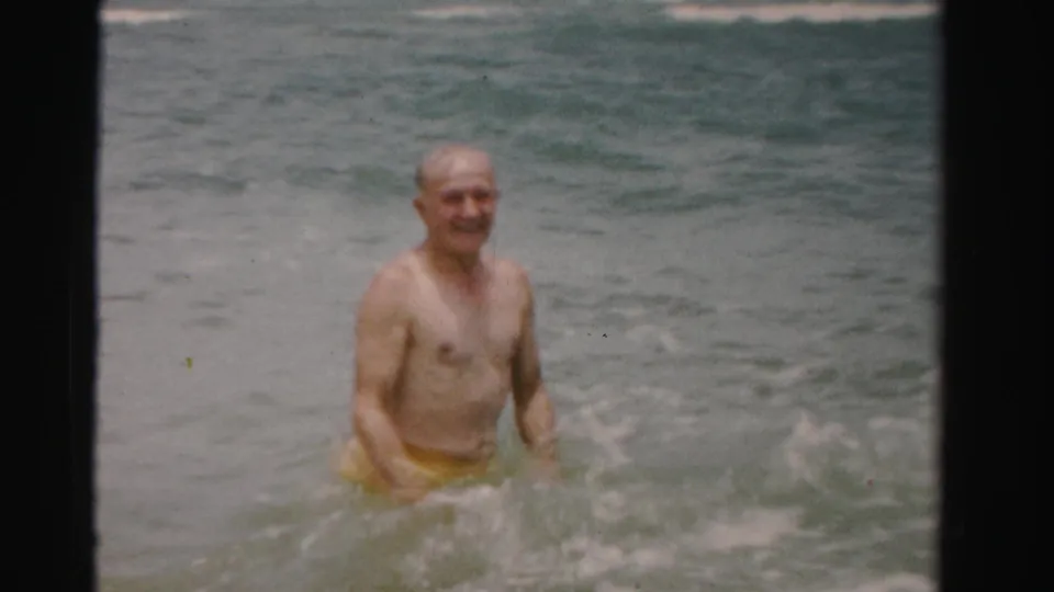 Older man in yellow swim trunks turns slightly as shallow waves move around him at a Florida beach.