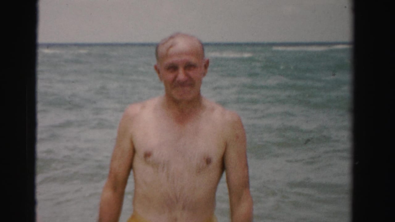 Elderly shirtless man standing in ocean surf and facing the camera.