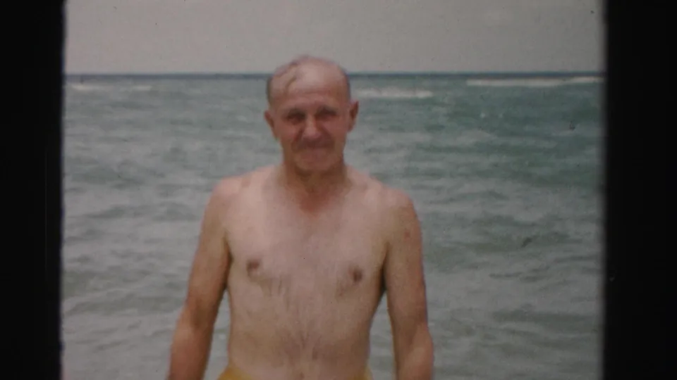In 1955 Dade County beach footage, an elderly shirtless man stands in shallow ocean waves and looks toward the camera.