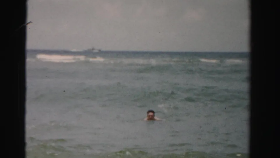 A distant swimmer remains visible above the rolling surf, framed by open water and a low horizon in the 1955 beach clip.