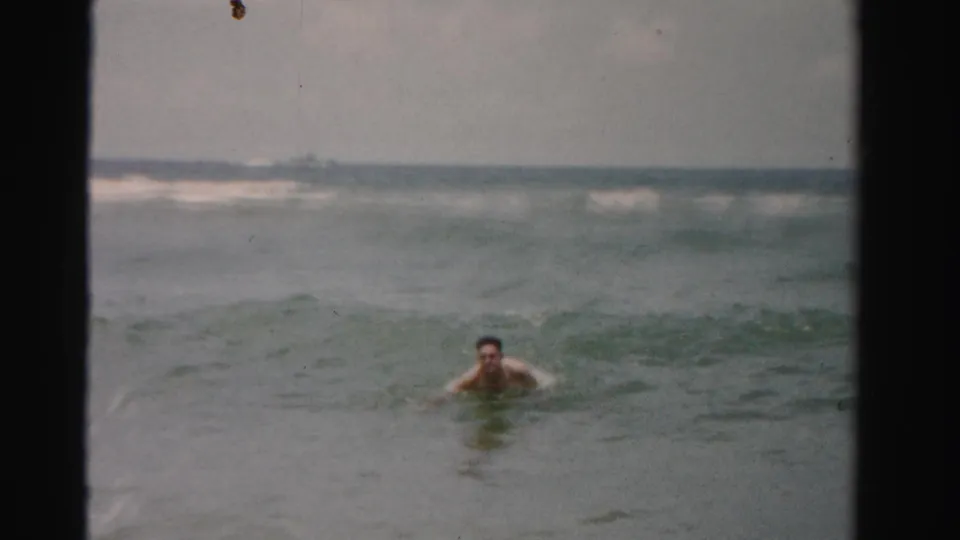 1955 Dade County home-movie frame showing an adult man facing the camera while swimming in rolling ocean surf.
