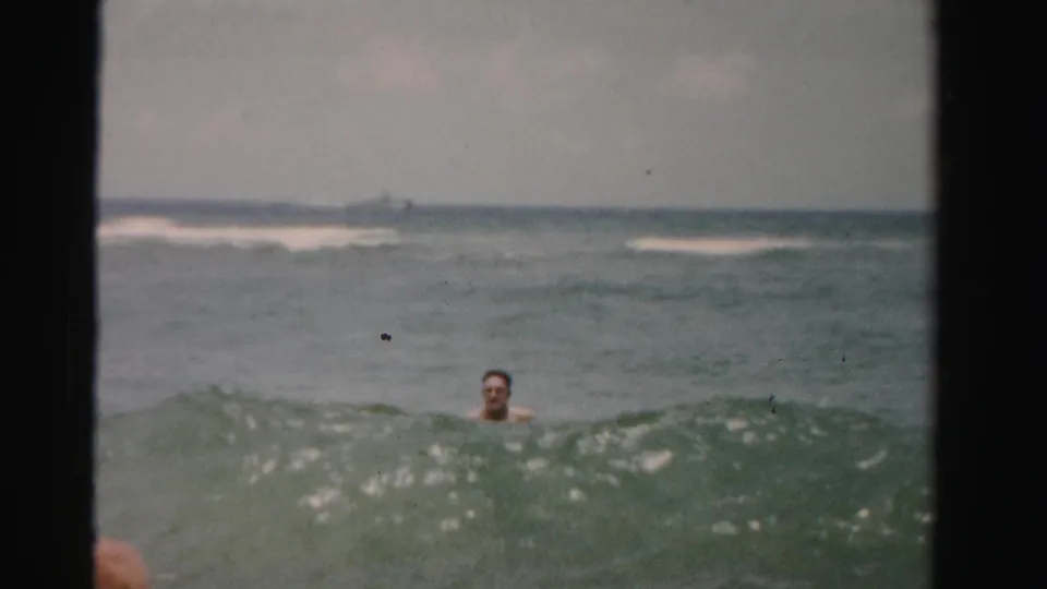 Adult swimmer in Dade County surf, partly obscured by a wave, with a faint distant boat or shoreline form on the horizon.